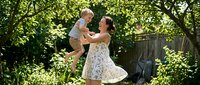 Young Caucasian mother in floral dress playing with toddler son in sunny backyard garden. Happy family bonding moment outdoors among green trees and foliage.