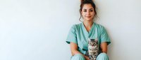 Young female veterinarian in mint green scrubs holding adorable tabby cat while sitting against white wall, representing healthcare and animal care services.