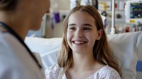 Young female patient with bright smile in hospital bed, wearing white gown, receiving medical care. Positive healthcare experience shown through genuine expression.
