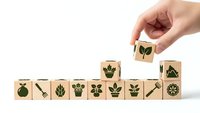 Hand placing wooden cube with leaf symbol on stack of eco-friendly blocks featuring nature icons on white background for sustainable business concepts.