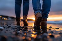 Couple walking on wet beach at sunset, close-up view of boots in shallow water with golden light reflections on pebbles creating romantic atmosphere.
