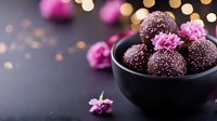 Dark chocolate truffles decorated with pink carnation flowers in black ceramic bowl against blurred bokeh lights background, romantic dessert setting.