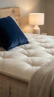 Cozy bedroom interior with quilted white mattress, navy blue pillow, wooden headboard, and warm bedside lamp creating relaxing ambiance for sleep and rest.