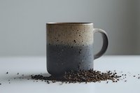 Minimalist ceramic mug in two-tone gray and beige design with speckled texture, surrounded by scattered loose tea leaves on white surface, artistic food photography.