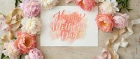 Happy Mother's Day greeting card surrounded by pink peonies, ranunculus flowers and ribbon on rustic wooden background for holiday celebration.