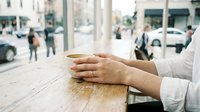 Woman's hands holding a warm coffee cup at a wooden cafe counter with a blurred city street in the background.