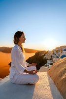 Young woman meditating in lotus pose at sunset overlooking Mediterranean village with white buildings and blue domes during golden hour wellness retreat.