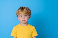 Young caucasian boy with blonde hair wearing bright yellow t-shirt making sad pouty expression against vibrant blue background, studio portrait showing childhood emotions.