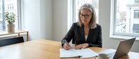 Mature businesswoman with glasses writing notes at office desk with laptop and documents in bright modern workspace.