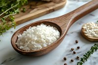 Raw white rice grains in wooden spoon on marble surface with fresh herbs and peppercorns, natural food styling for cooking and culinary concepts.