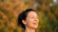 Happy middle-aged woman with curly hair laughing outdoors in autumn park with golden foliage background for wellness and lifestyle content.