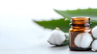 Brown glass essential oil bottle with fresh cotton flowers and green leaves on light background, natural organic skincare concept.