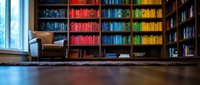 Cozy library interior with colorful rainbow organized bookshelves, leather armchair and wooden floor creating perfect reading atmosphere for study.