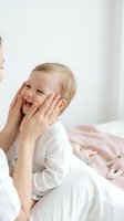 Happy blonde toddler boy laughing with mother's hands gently touching his face in bright bedroom. Joyful family bonding moment in white pajamas on bed.