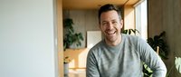 Happy middle-aged Caucasian man in gray sweater laughing in modern kitchen with plants and natural lighting for lifestyle and wellness content.