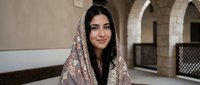 Young Middle Eastern woman wearing traditional embroidered headscarf smiling warmly in historic stone archway setting for cultural lifestyle photography.