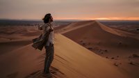 A young woman with curly dark hair, wearing casual travel clothing and a backpack, stands on a towering sand dune at sunset, gazing across vast desert landscape with golden light illuminating the endless sandy terrain.