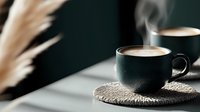 Two steaming cups of coffee in dark mugs sit on woven coasters against a dark background with dried pampas grass, creating a cozy morning or afternoon beverage scene.