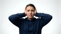 Young Caucasian woman in navy hoodie covering ears with hands showing stressed overwhelmed expression on white background for mental health concepts.