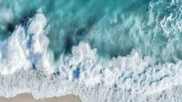Aerial view of ocean waves crashing on sandy beach with white foam patterns creating abstract natural texture in turquoise and teal colors. Perfect for wall art.