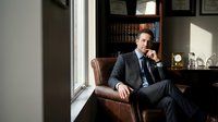 Professional businessman in suit sitting thoughtfully in office with bookshelf and natural window light for corporate and business concepts.