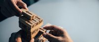 Hands collaborating to build a wooden block tower with business icons, symbolizing teamwork, strategy, and corporate growth.