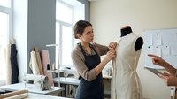 Young female fashion designer adjusting white garment on dress form mannequin in bright modern atelier workspace with sewing equipment and fabric rolls.