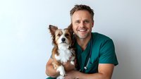 Smiling male veterinarian in green scrubs holding small brown and white dog in clinic setting for pet healthcare and animal medical services.