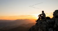 Man sitting on rocky cliff watching birds fly across golden sunset sky over mountain landscape for meditation and outdoor adventure concepts.
