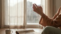 Young woman practicing morning meditation with raised hands in sunlit room by window for mindfulness and spiritual wellness routine.