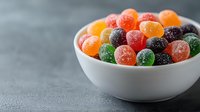 Colorful sugar-coated gummy candies in white ceramic bowl on gray background. Sweet fruit-flavored jelly drops with crystallized coating for confectionery design.