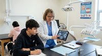 Female dentist in white coat consulting with young male patient using digital tablet in modern dental office with equipment and educational posters.