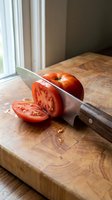 Fresh ripe tomato being sliced on wooden cutting board with chef knife in bright kitchen with natural window light, food preparation.