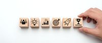 Hand arranging wooden blocks with business success icons showing teamwork, innovation, growth, target, rocket and trophy symbols.