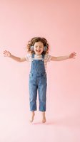 Happy young girl with curly hair wearing headphones and denim overalls dancing with arms outstretched on pink background isolated for design use.
