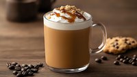 Caramel latte topped with whipped cream and drizzle in glass mug on wooden table with coffee beans and chocolate chip cookie, warm brown tones.