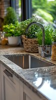 Modern kitchen interior with stainless steel sink and chrome faucet on granite countertop, decorated with fresh herbs in wicker baskets and ceramic pots near window.