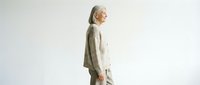 Senior woman with long gray hair wearing a beige knit cardigan and light pants, standing in profile against white background for fashion and lifestyle content.
