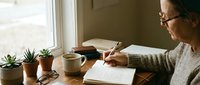 Young Caucasian woman with glasses writing in notebook at wooden desk by window with coffee cup and small potted plants in cozy home office workspace.