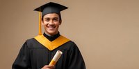 Young Asian male graduate in black gown with yellow trim holding diploma against neutral beige background, celebrating academic achievement.