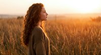 Young woman with curly hair standing in golden wheat field during sunset, peaceful countryside moment with warm lighting for wellness and lifestyle content.