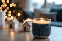 Glowing scented candle in geometric pattern holder with decorative ceramic houses and soft bokeh lights creating cozy winter atmosphere on wooden table.
