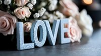 Decorative blue love text with delicate pink roses and baby breath flowers on dark background, romantic wedding or valentines day composition with soft bokeh.