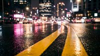 Wet city street at night with glowing yellow road markings, bokeh lights, and urban skyline reflecting on rain-soaked asphalt.