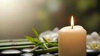 Lit candle with bamboo leaves and white orchid flowers on spa stones creating peaceful zen atmosphere for relaxation and meditation wellness treatments.