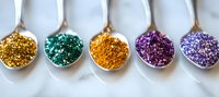 Metallic glitter powder in silver spoons arranged in row showing gold, emerald, copper, purple and lavender colors on white background. Macro photography.