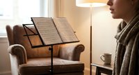 Young woman musician reading sheet music on stand in cozy living room with warm lighting, practicing at home for performance or lesson preparation.