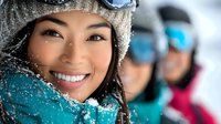Young Asian woman smiling brightly in winter sports gear with snow on face and knit hat, blurred skiers in background. Active lifestyle and winter recreation concept.
