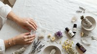 Woman hands creating natural aromatherapy blend with essential oils, dried herbs, crystals and botanical ingredients on linen surface for wellness.