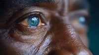 Close up macro shot of bright blue eye with dark skin around, showing intricate iris details and natural eye reflection in dramatic lighting.